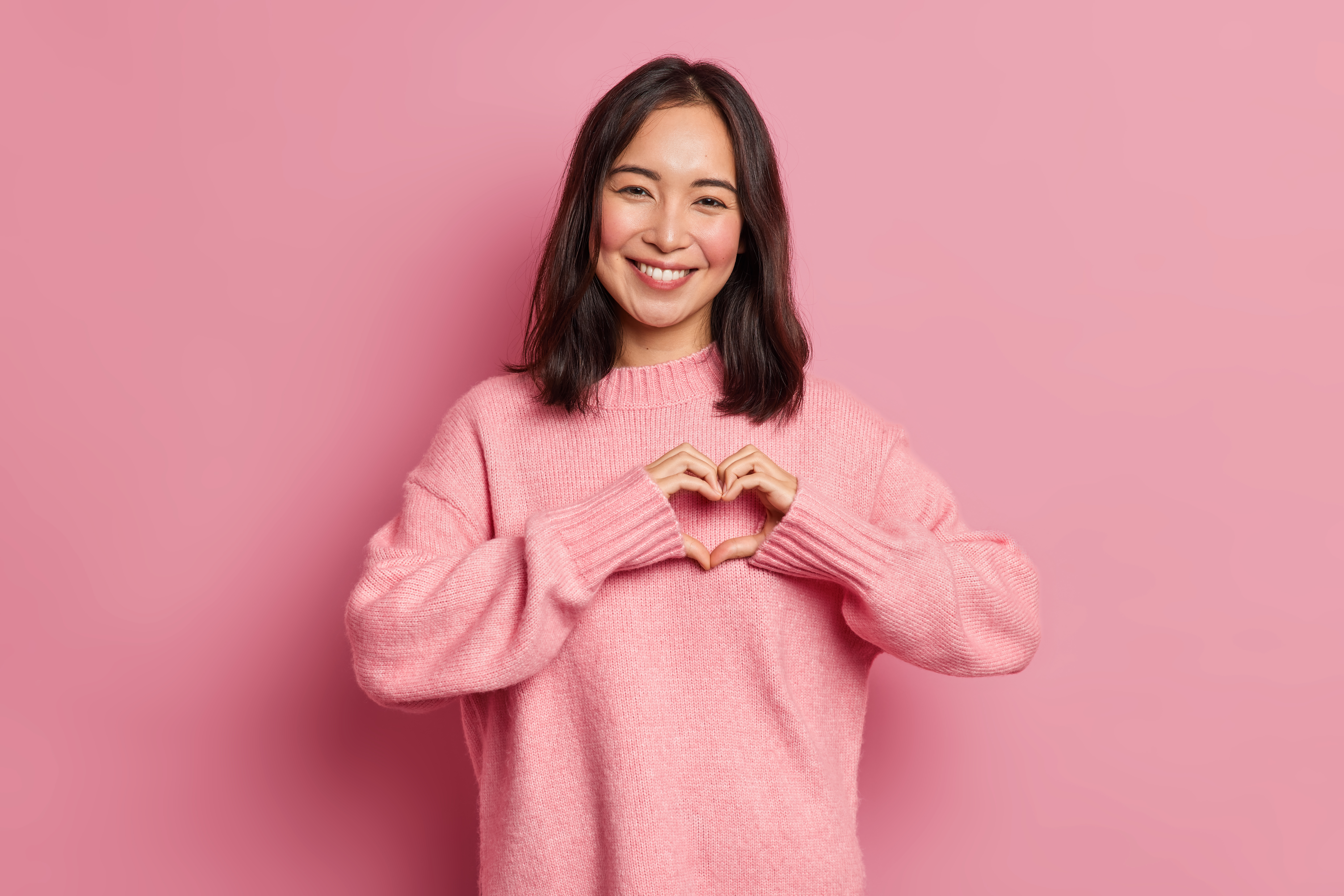 Young brunette woman smiles happily for the camera while holding her hands in the shape of a heart in front of a pink background.