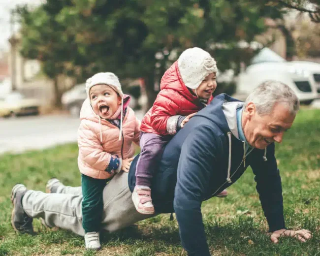 grandfather with two young grandchildren sitting on his back while he performs push up exercises