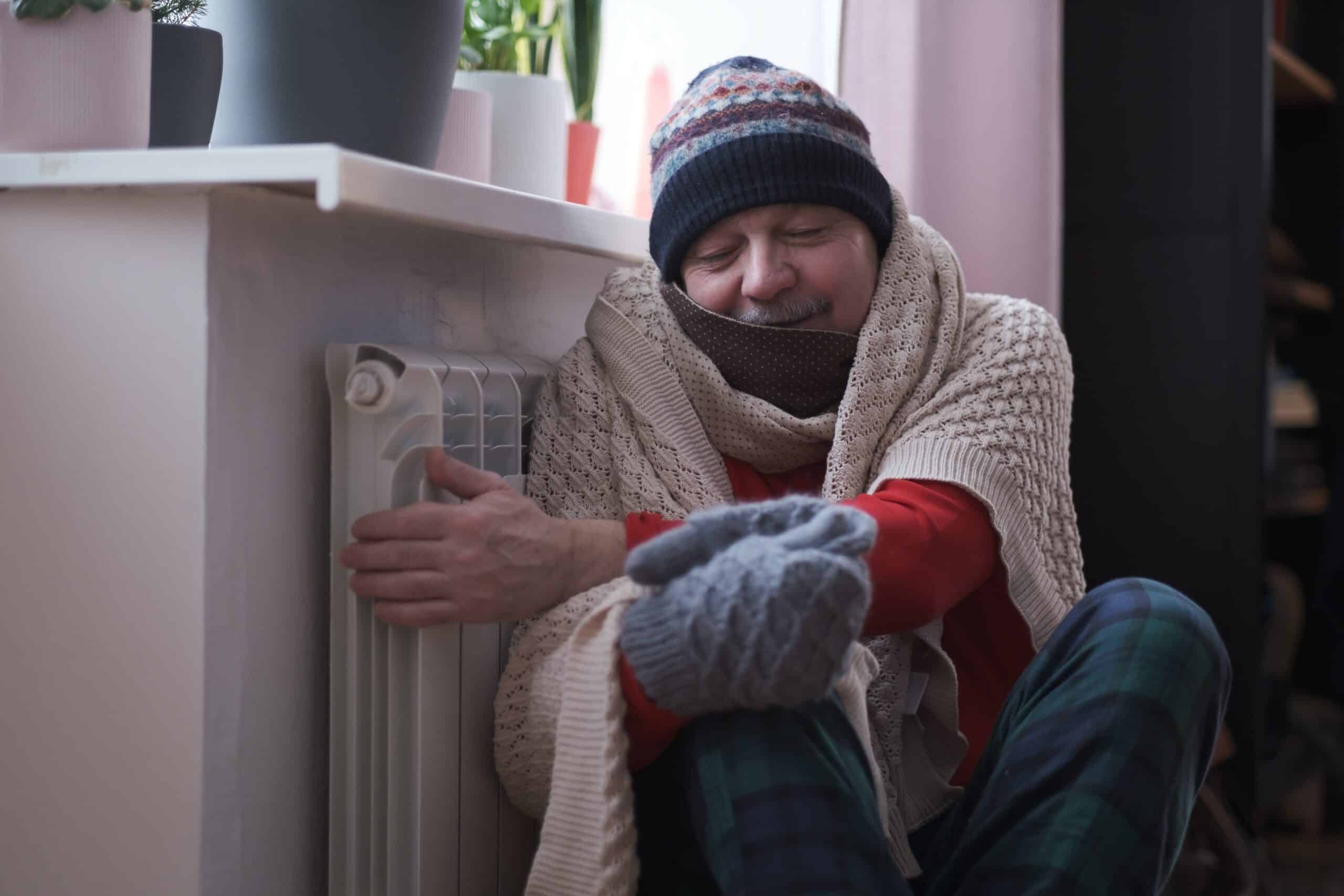 Man sits on floor next to his furnace while wearing winter clothing to try and warm himself.